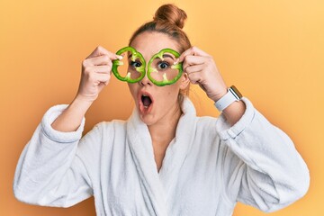 Poster - Young blonde woman wearing bathrobe holding green pepper over eyes afraid and shocked with surprise and amazed expression, fear and excited face.