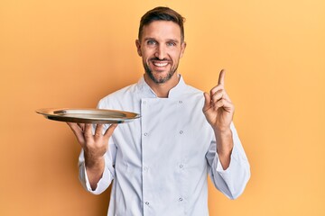 Wall Mural - Handsome man with beard wearing chef uniform holding silver tray smiling happy pointing with hand and finger to the side