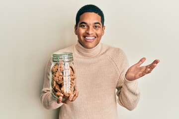 Young handsome hispanic man holding jar of chocolate chips cookies celebrating achievement with happy smile and winner expression with raised hand