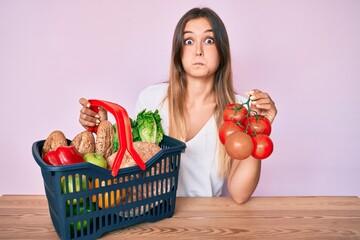 Beautiful caucasian woman holding supermarket shopping basket and tomatoes puffing cheeks with funny face. mouth inflated with air, catching air.