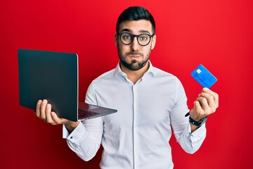 Canvas Print - Young hispanic man wearing business style holding laptop and credit card puffing cheeks with funny face. mouth inflated with air, catching air.