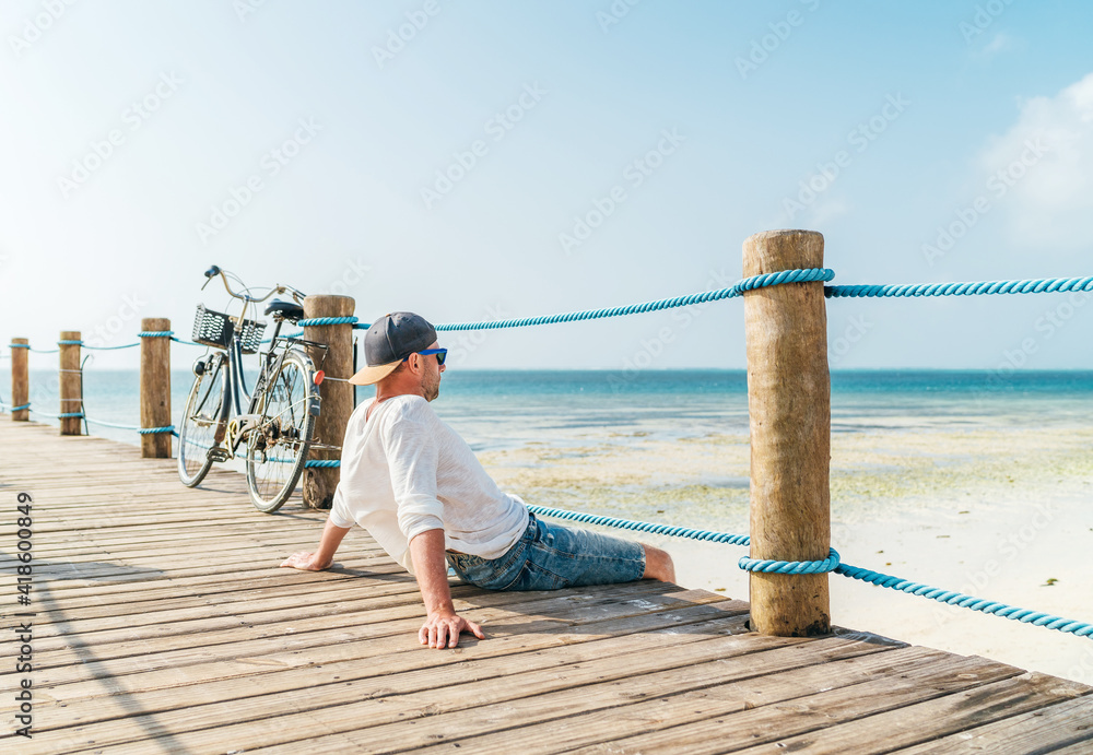 Portrait of relaxing man dressed in light summer clothes and sunglasses sitting and enjoying time and beach view on wooden pier.Careless vacation in tropical countries concept image. Zanzibar,Tanzania - obrazy, fototapety, plakaty 