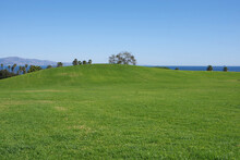 Panoramic Landscape View With A Large Grass Area With A Hill In Front And Trees And Palms And The Pacific Ocean At Santa Barbara And Mountains Behind Under Blue Sky