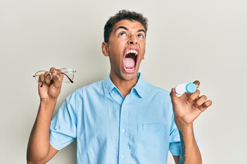 Sticker - Young handsome african american man holding glasses and contact lenses angry and mad screaming frustrated and furious, shouting with anger looking up.