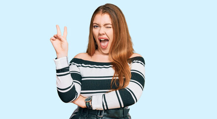 Wall Mural - Young beautiful redhead woman wearing elegant clothes smiling with happy face winking at the camera doing victory sign. number two.