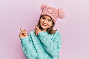 Little beautiful girl wearing wool sweater and cute winter hat smiling and looking at the camera pointing with two hands and fingers to the side.