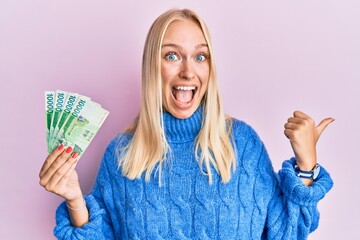 Wall Mural - Young blonde girl holding 10000 south korean won banknotes pointing thumb up to the side smiling happy with open mouth