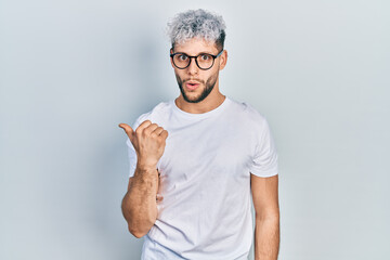 Young hispanic man with modern dyed hair wearing white t shirt and glasses surprised pointing with hand finger to the side, open mouth amazed expression.