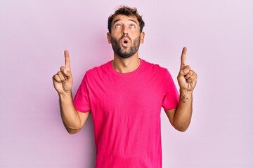 Handsome man with beard wearing casual pink tshirt over pink background amazed and surprised looking up and pointing with fingers and raised arms.