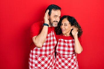 Poster - Middle age couple of hispanic woman and man wearing cook apron smiling with hand over ear listening an hearing to rumor or gossip. deafness concept.