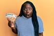 Young black woman with braids holding bunch of 50 euro banknotes scared and amazed with open mouth for surprise, disbelief face
