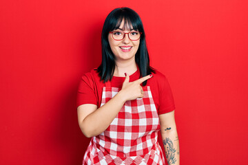 Sticker - Young hispanic woman wearing cook apron and glasses cheerful with a smile of face pointing with hand and finger up to the side with happy and natural expression on face