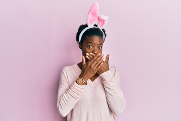 Wall Mural - Young african american girl wearing cute easter bunny ears shocked covering mouth with hands for mistake. secret concept.