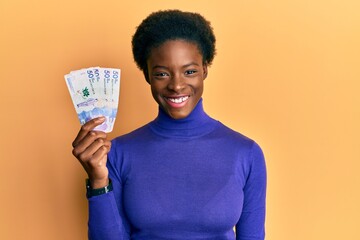 Poster - Young african american girl wearing doctor uniform holding 50 colombian pesos looking positive and happy standing and smiling with a confident smile showing teeth