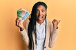 © Krakenimages.com - African american woman holding south african rand banknotes pointing thumb up to the side smiling happy with open mouth