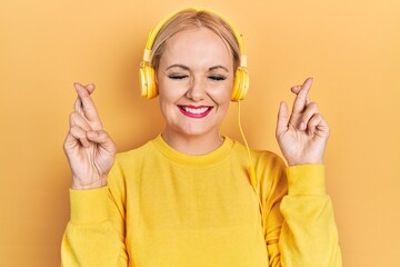 Canvas Print - Young blonde woman listening to music using headphones gesturing finger crossed smiling with hope and eyes closed. luck and superstitious concept.