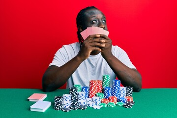 Canvas Print - Handsome young black man playing gambling poker covering face with cards smiling looking to the side and staring away thinking.