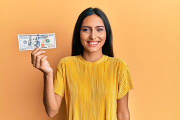 Canvas Print - Young brunette woman holding 100 dollars banknote looking positive and happy standing and smiling with a confident smile showing teeth