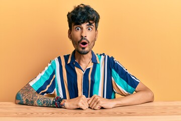 Canvas Print - Young hispanic man wearing casual clothes sitting on the table scared and amazed with open mouth for surprise, disbelief face