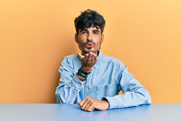 Poster - Young hispanic man wearing casual clothes sitting on the table looking at the camera blowing a kiss with hand on air being lovely and sexy. love expression.