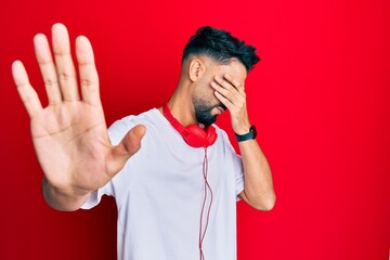 Young man with beard listening to music using headphones covering eyes with hands and doing stop gesture with sad and fear expression. embarrassed and negative concept.