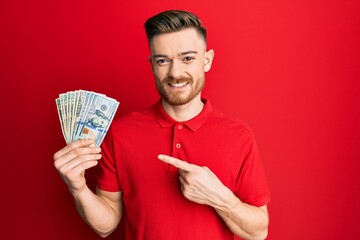 Young redhead man holding dollars smiling happy pointing with hand and finger