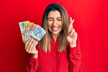 Poster - Beautiful brunette woman holding australian dollars gesturing finger crossed smiling with hope and eyes closed. luck and superstitious concept.