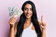 © Krakenimages.com - Beautiful hispanic woman holding dollars smiling with an idea or question pointing finger with happy face, number one
