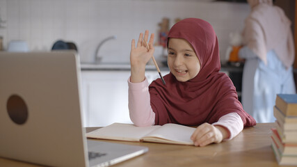 The little female student wearing a headscarf is watching the lessons on the internet with a computer video call and working from home. She asks questions to her teacher and takes notes. 