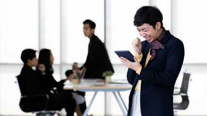 Portrait of LGBTQ transgender man office employee in casual suit standing using and holding tablet computer with cheerful and happy gesture with team colleagues blur in background