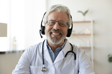 Wall Mural - Headshot portrait of smiling mature Caucasian male doctor in headphones talk on video call in hospital. Profile picture of happy senior man GP or therapist in earphones consult patient on webcam.