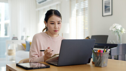 Candid young attractive asian female employee wear earphone listen to digital online course development program in quarantine social distance at home reskill upskill school class for career path.