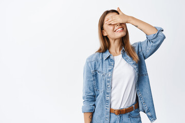 Wall Mural - Portrait of smiling beautiful woman waiting for surprise while eyes covered with hand, anticipating something good, standing against white background