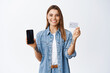 © Cookie Studio - Online shopping. Portrait of smiling blond girl recommending shop, showing empty smartphone screen and plastic credit card, standing over white background