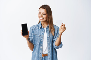 Wall Mural - Online shopping. Portrait of young woman making money on app, showing empty smartphone screen and plastic credit card smiling, looking at mobile application, white background