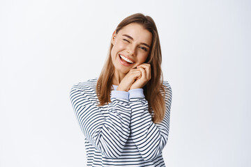 Wall Mural - Cheeky young woman with blond hair, holding hands near natural face with light make up, winking and smiling at camera, standing over white background