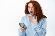 Shocked and startled redhead girl scream while reading message on smartphone, staring at mobile screen with surprised face, standing against white background