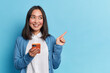 © wayhome.studio  - Beautiful Asian woman uses smartphone app sends messages in social media chat points away on copy space wears casual jumper isolated over blue background shows copy space for your advertisement