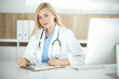 © Iryna - Woman-doctor at work while sitting at the desk in hospital or clinic. Blonde cheerful physician filling up medication history record form