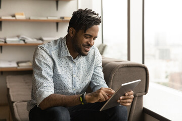 Close up positive African American young man using tablet, having fun, sitting on couch at home, browsing apps, chatting, spending leisure time with gadget, satisfied customer shopping online