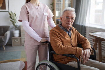 Wall Mural - Cropped portrait of senior man in wheelchair looking at camera with unrecognizable nurse assisting him, copy space