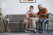 Full Length Portrait Of Young Female Nurse Assisting Senior Man In Wheelchair Using Digital Tablet At Retirement Home, Both Wearing Masks, Copy Space