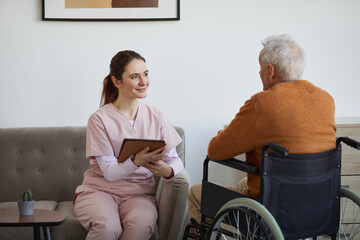 Wall Mural - Portrait of smiling female nurse talking to senior man in wheelchair and using digital tablet at retirement home, copy space