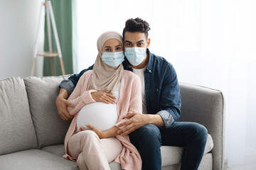 Wall Mural - Young pregnant muslim couple wearing medical masks sitting on couch at home