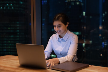 Close up smiling confident Indian businesswoman using laptop at night in office, working, finishing project at late hours, deadline, looking at computer screen, sitting at desk near panoramic window