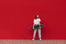 Horizontal Full-length Portrait Of A Stylish Female Skater Holding Her Skateboard And Standing On A City Street In Front Of A Red Wall With A Copy Space.