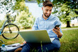 © baranq - Young businessman checking phone while sitting in the park with bicycle and laptop