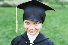 Back To Elementary School Concept. Little Girl Goes To First Grade. Ceremony Of Graduating. Black Gown, Academic Cap With Tassels. Party With Parents, Teachers In Park, Yard. Last Day, End Of Year