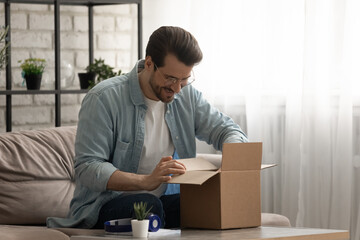 Present from distant friend. Happy millennial man unpack open box with birthday gift surprise received by mail. Smiling young guy has pleasure to receive package with consumer goods purchased online