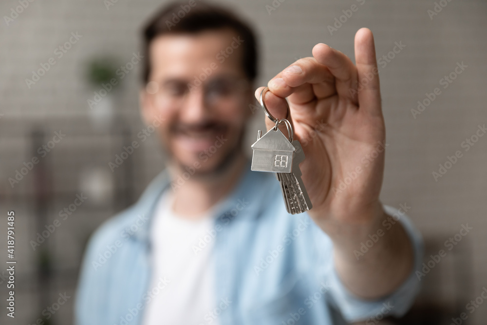 Satisfied homeowner. Blurred portrait of happy young man buyer renter of new modern home apartment holding key demonstrating wellbeing wealth celebrate achievement. Focus on hand with keys of dwelling - obrazy, fototapety, plakaty 